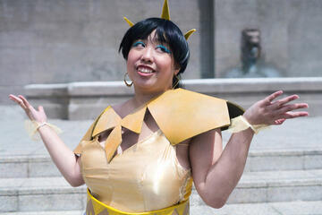 Pōhutukaryl Cosplay as a human version of Sagira from Destiny: gold dress, spikes in hair, shoulder pauldrons